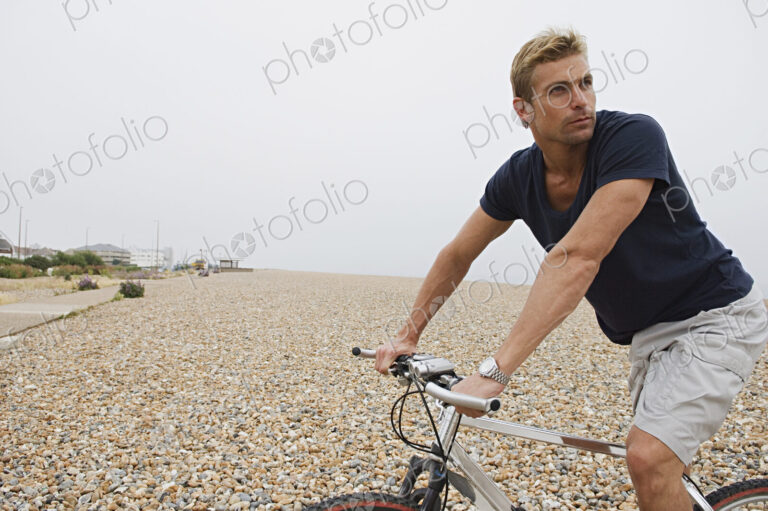 A man riding a bicycle on a shingle beach.