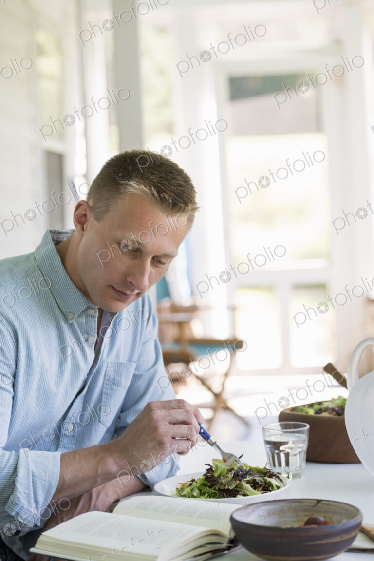 A man seated at a cafe table eating a salad and reading a book.