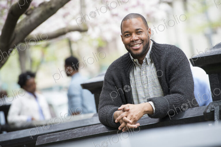 A man seated at a table outdoors, two people in the background.