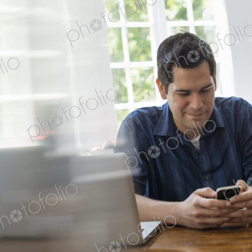 A man seated at a table with a laptop, checking his smart phone. A man seated at a table with a laptop, checking his smart phone.