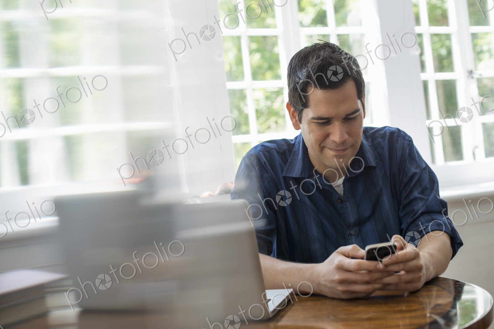 A man seated at a table with a laptop, checking his smart phone.