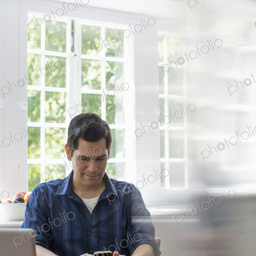 A man seated at a table with a laptop, checking his smart phone. A man seated at a table with a laptop, checking his smart phone.