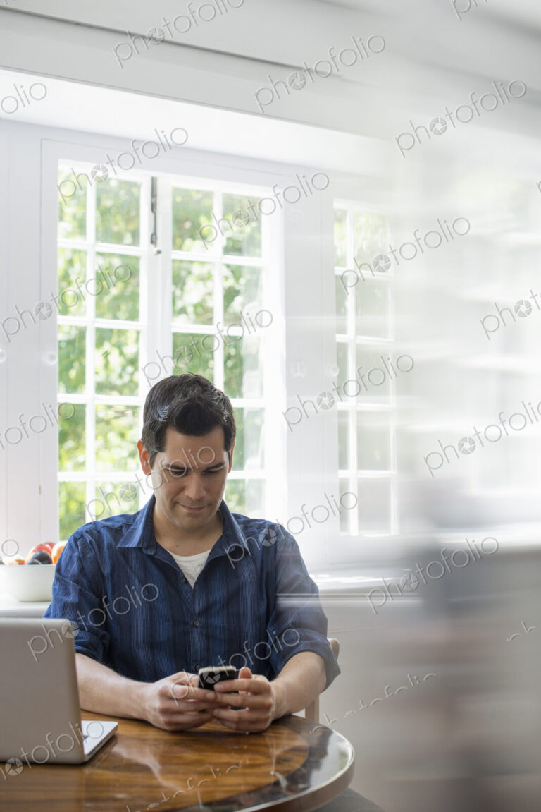 A man seated at a table with a laptop, checking his smart phone.