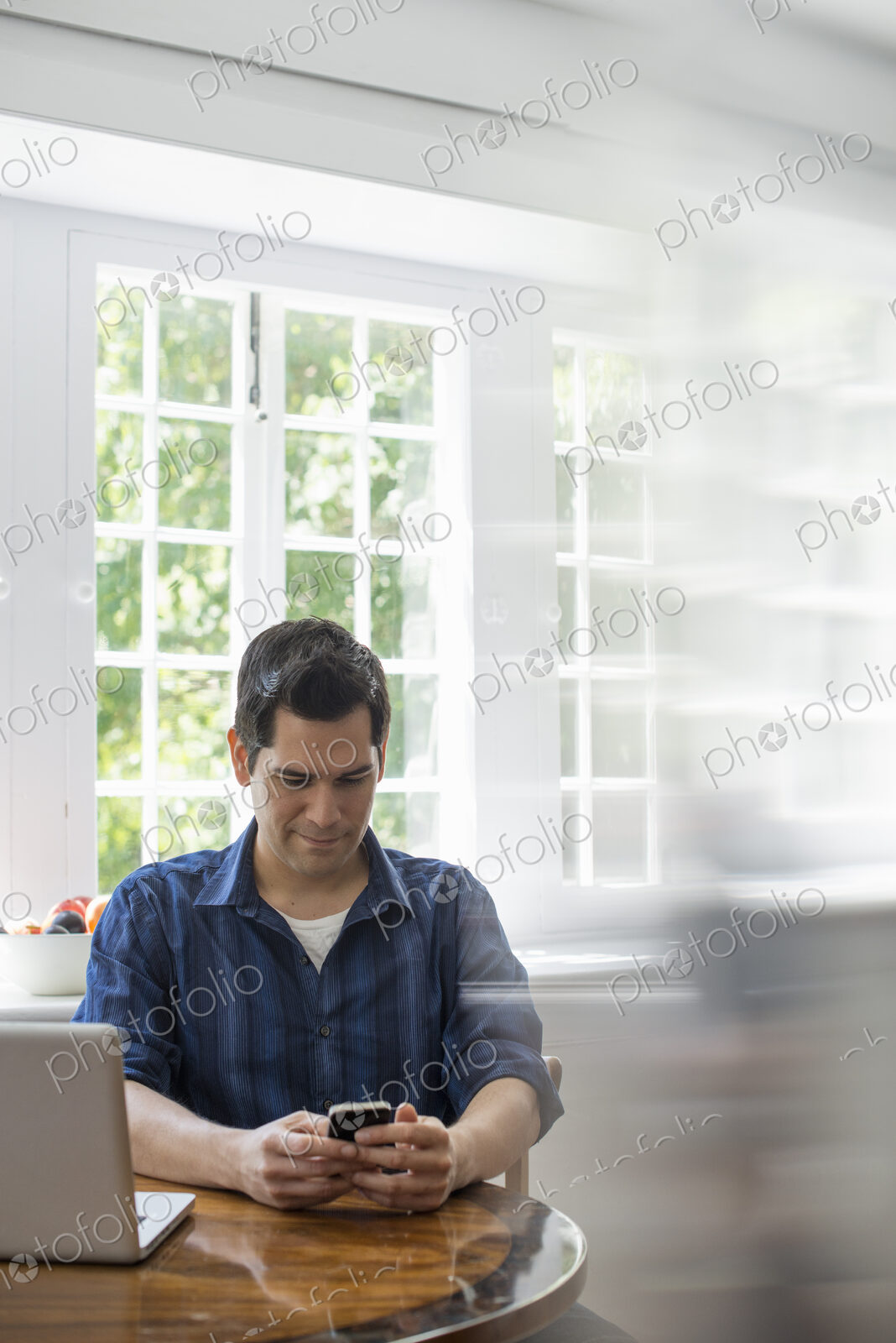 A man seated at a table with a laptop, checking his smart phone.