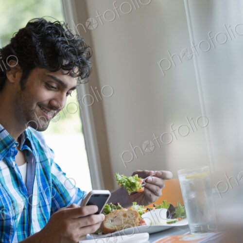 A man seated checking his phone, and eating in a cafe. A man seated checking his phone, and eating in a cafe.