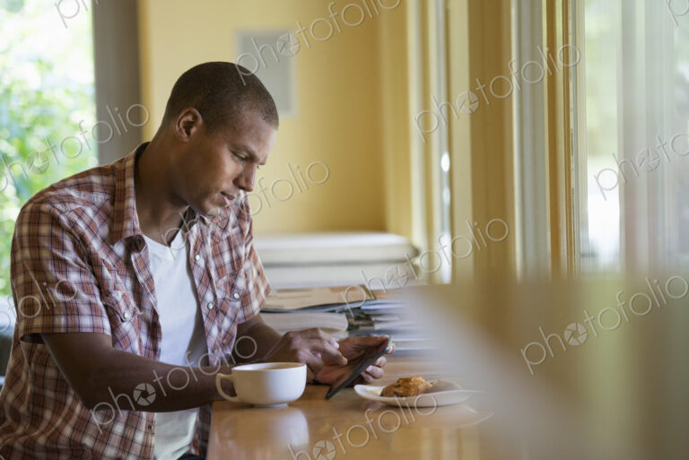 A man seated having a cup of coffee, holding a digital tablet.