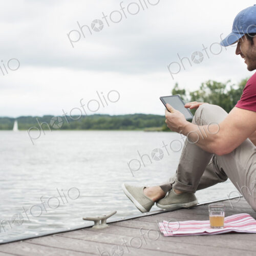 A man seated on a jetty by a lake, using a digital tablet. A man seated on a jetty by a lake, using a digital tablet.