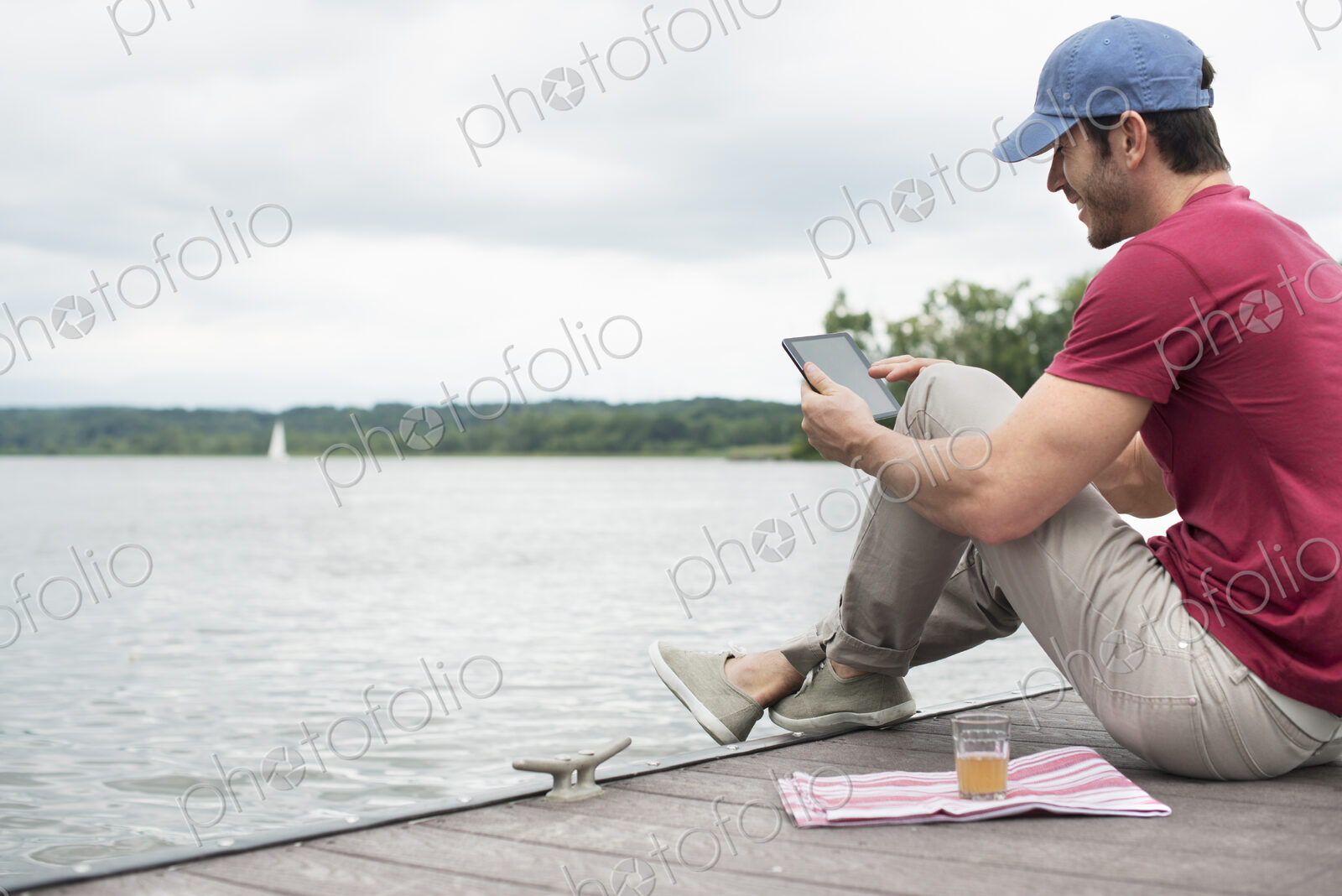 A man seated on a jetty by a lake, using a digital tablet.