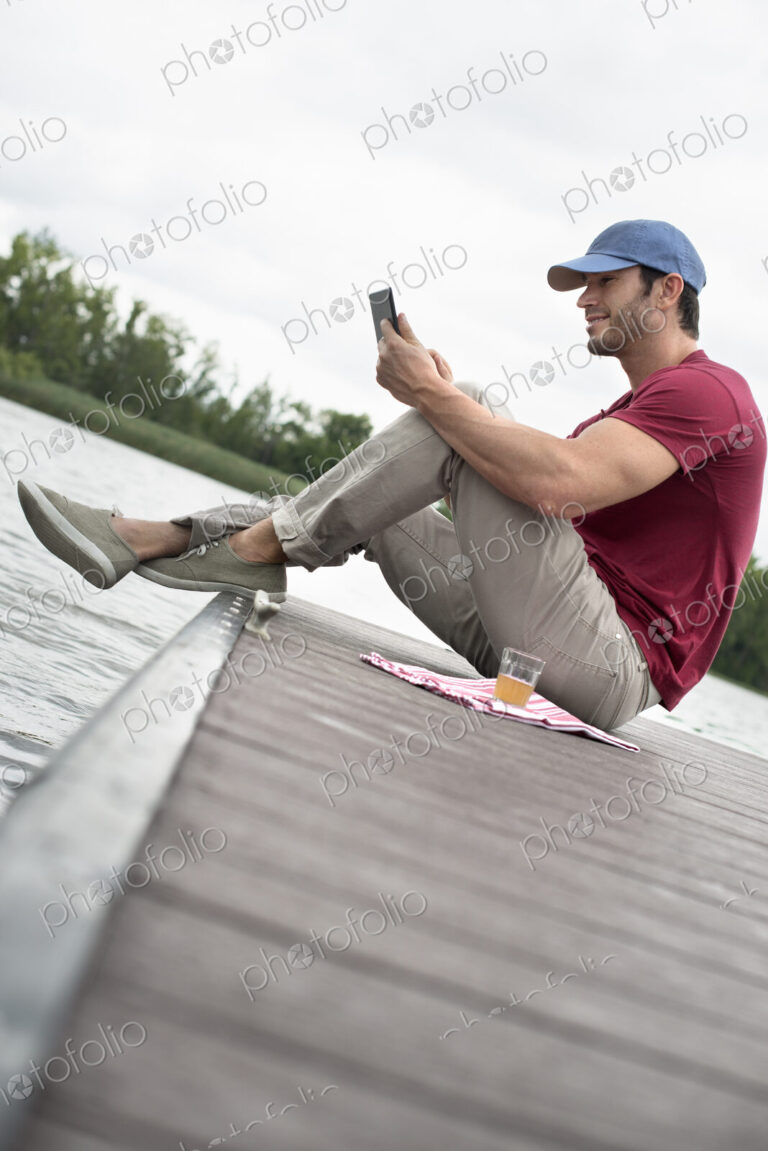 A man seated on a jetty by a lake, using a digital tablet.