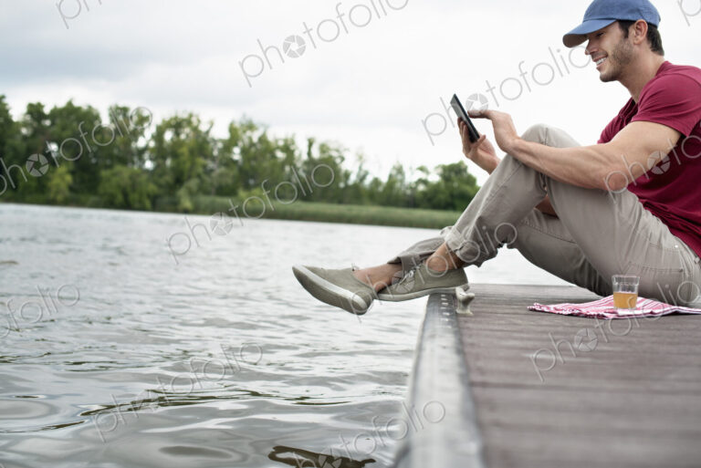 A man seated on a jetty by a lake, using a digital tablet.