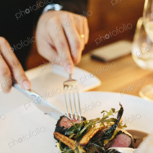 A man seated using a knife and fork eating meat and roasted vegetables.