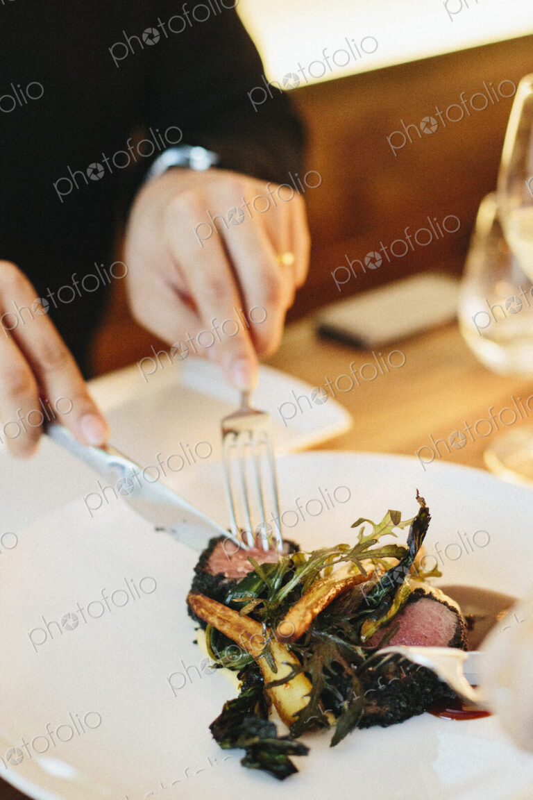 A man seated using a knife and fork eating meat and roasted vegetables.