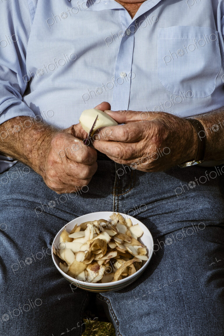 A man seated using a sharp knife to peel potatoes.