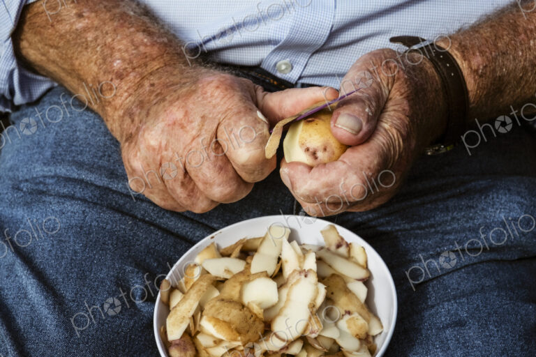 A man seated using a sharp knife to peel potatoes.