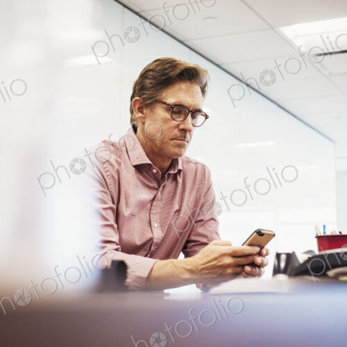 A man sitting in a classroom at a table and looking down at a cellphone. A man sitting in a classroom at a table and looking down at a cellphone.