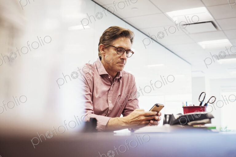 A man sitting in a classroom at a table and looking down at a cellphone.