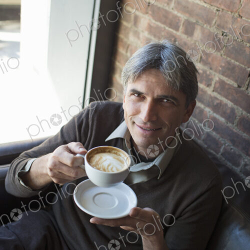 A man sitting in a coffee shop. Holding a cup of frothy cappuccino coffee.