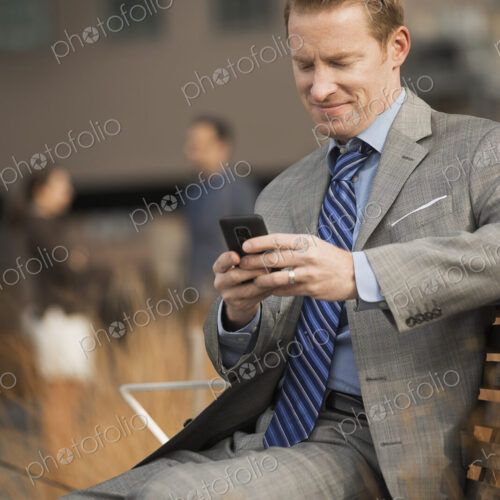 A man sitting on a bench outside a large building, looking at a cell phone screen or… A man sitting on a bench outside a large building, looking at a cell phone screen or…