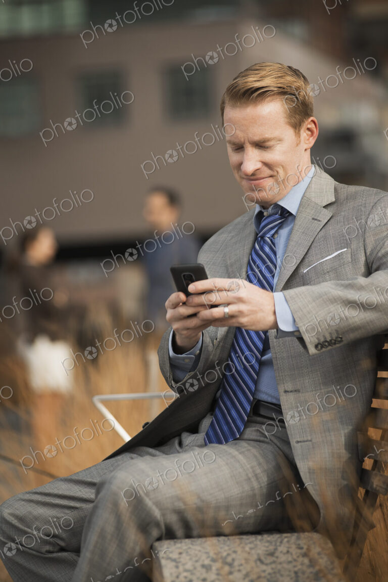 A man sitting on a bench outside a large building, looking at a cell phone screen or…