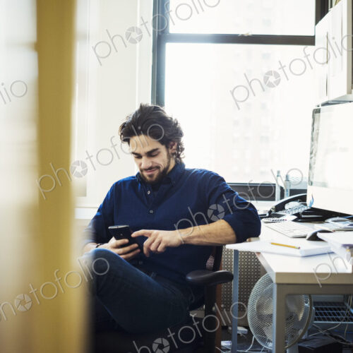 A man sitting on his own in an office checking his phone, seen through an open door. A man sitting on his own in an office checking his phone, seen through an open door.