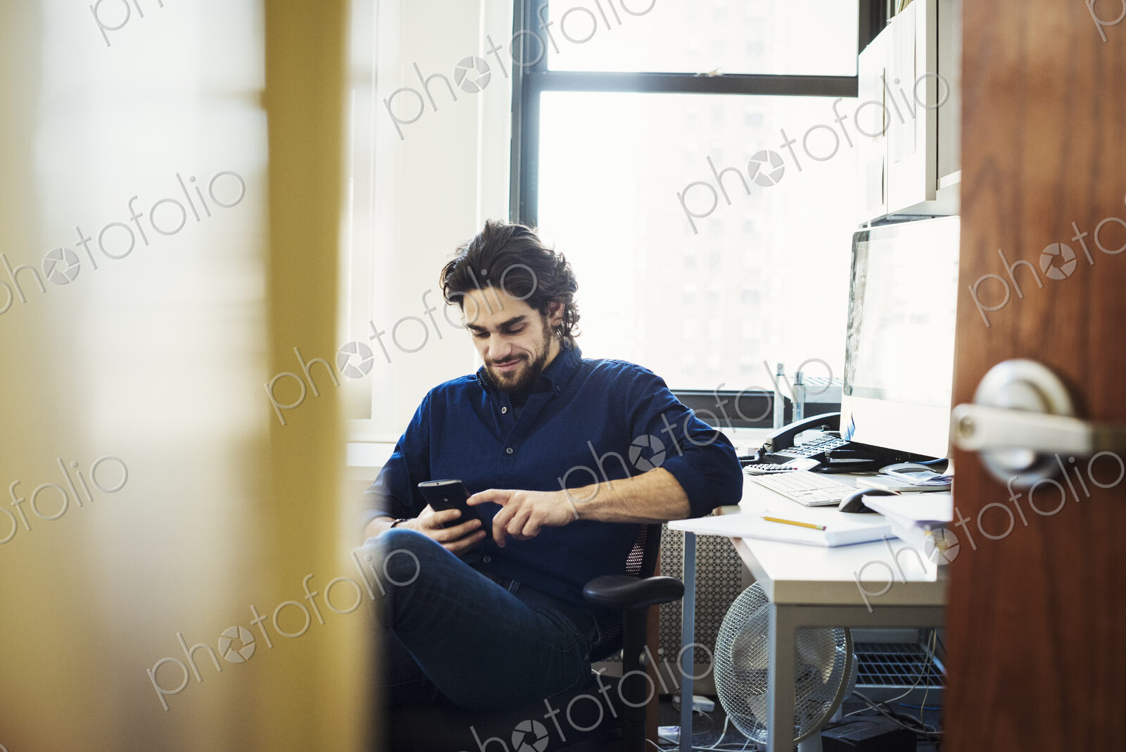 A man sitting on his own in an office checking his phone, seen through an open door.