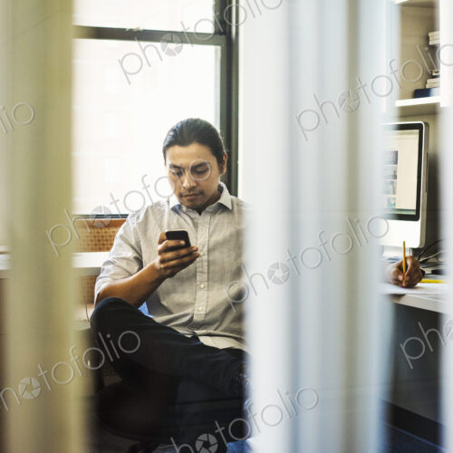 A man sitting on his own in an office checking his phone, seen through an open door.