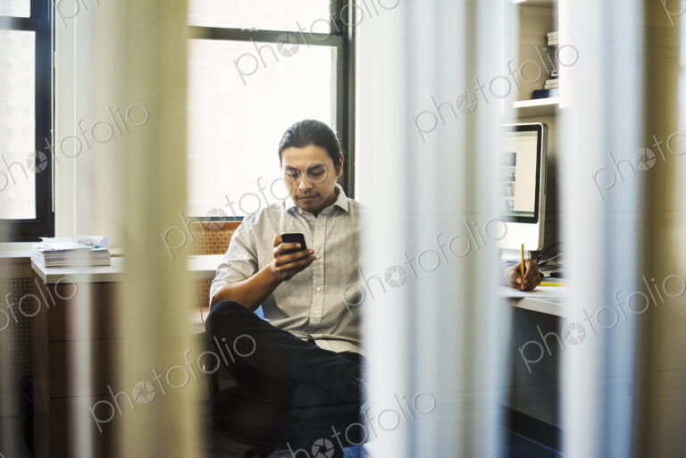 A man sitting on his own in an office checking his phone, seen through an open door.