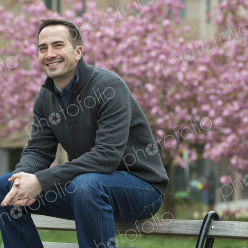 A man sitting on the back of a park bench, under the cherry blossom trees in the park.