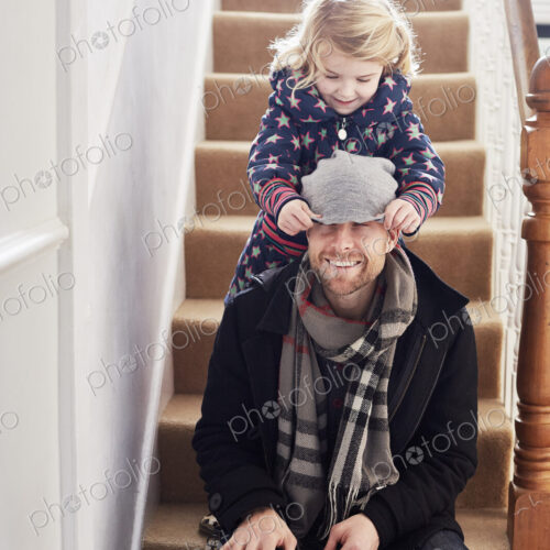 A man sitting on the stairs in a hallway, and a girl pulling his hat over his eyes.