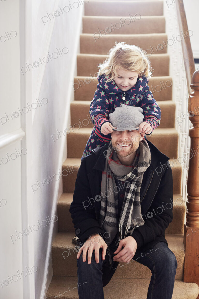 A man sitting on the stairs in a hallway, and a girl pulling his hat over his eyes.