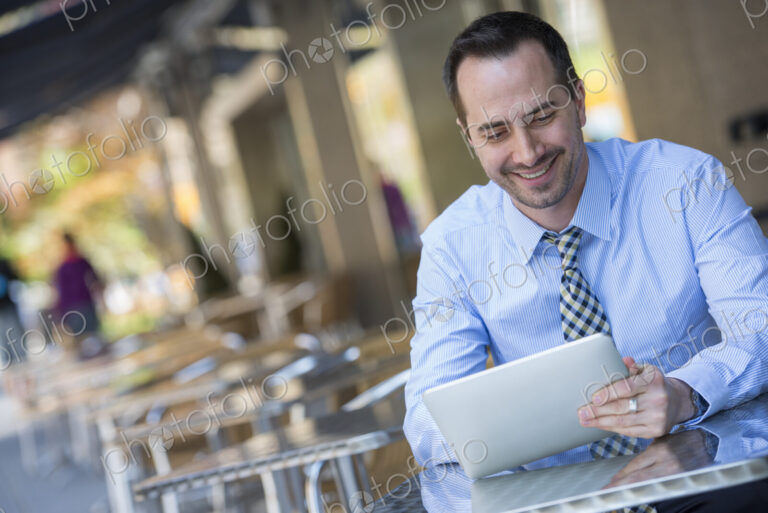 A man sitting outdoors at a cafe table using a digital tablet.