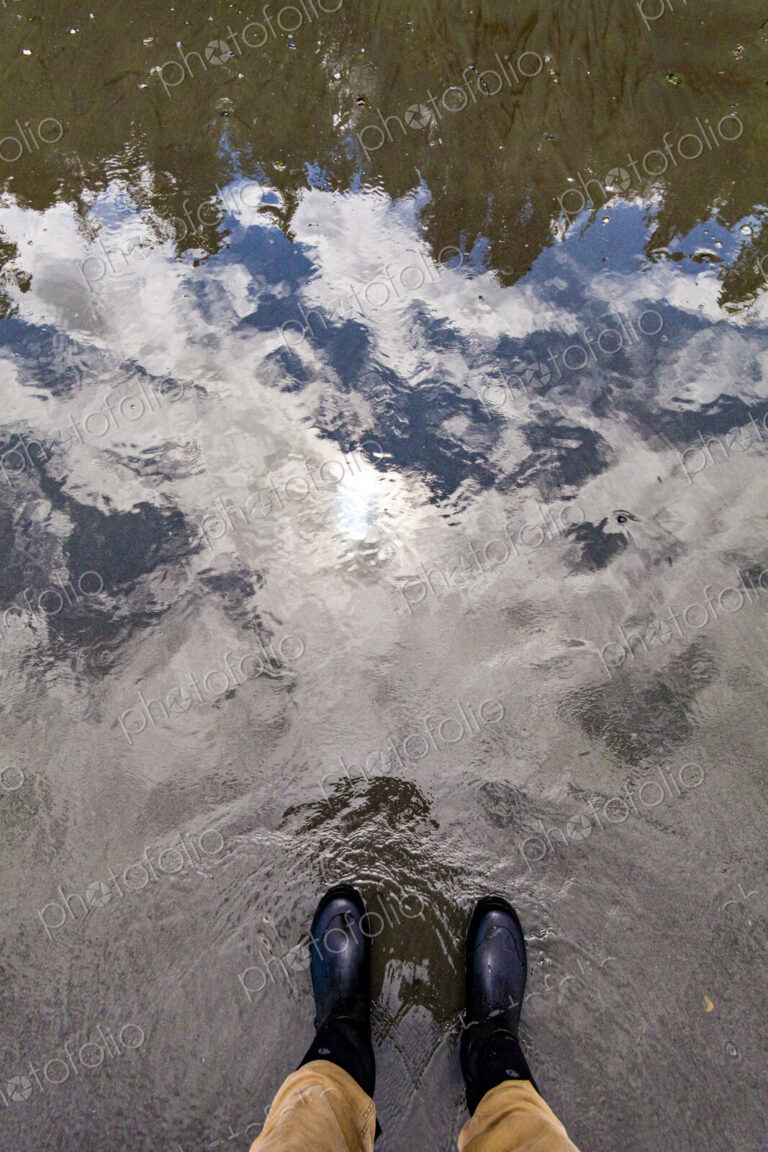 A man standing at the water's edge, a reflection of the forest and the sky in shallow…