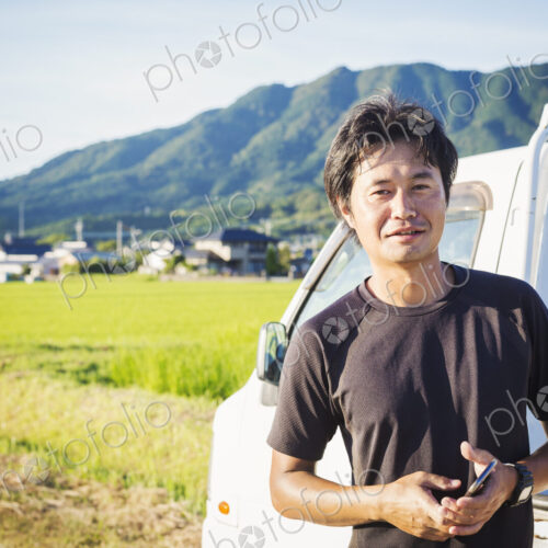 A man standing by a truck in an open landscape with rice paddies behind him, holding a… A man standing by a truck in an open landscape with rice paddies behind him, holding a…