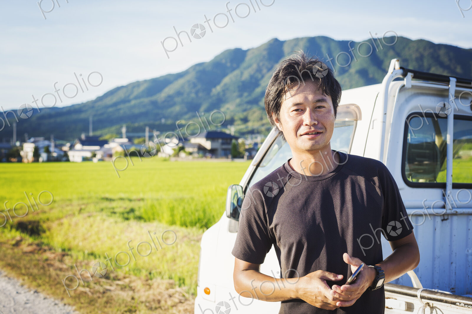 A man standing by a truck in an open landscape with rice paddies behind him, holding a…