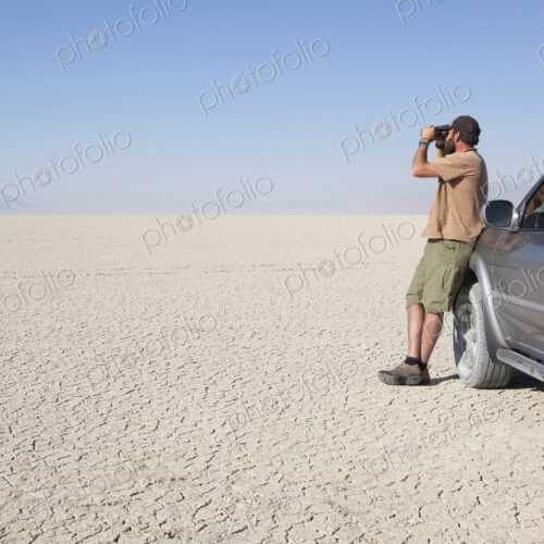 A man standing in a dry desert, looking through binoculars and leaning against a truck A man standing in a dry desert, looking through binoculars and leaning against a truck