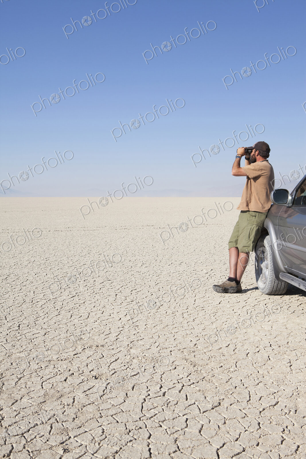 A man standing in a dry desert, looking through binoculars and leaning against a truck