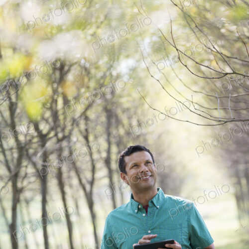 A man standing in an avenue of trees, holding a digital tablet. A man standing in an avenue of trees, holding a digital tablet.