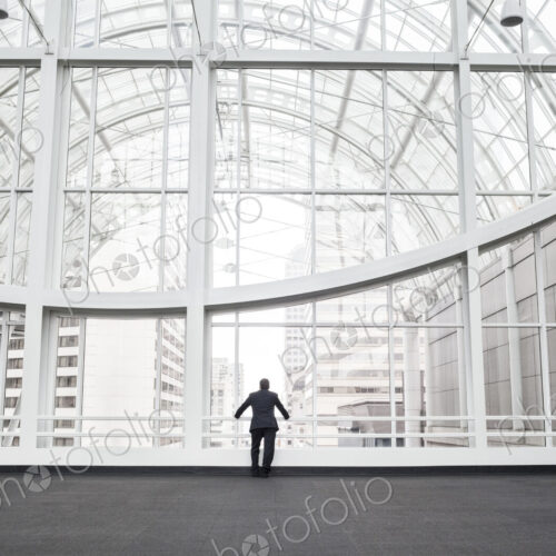 A man standing in an open space in a glass atrium in an office building, leaning on a…