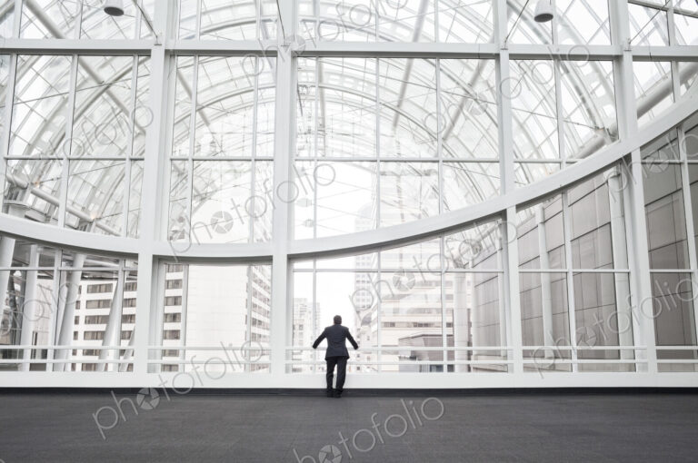 A man standing in an open space in a glass atrium in an office building, leaning on a…