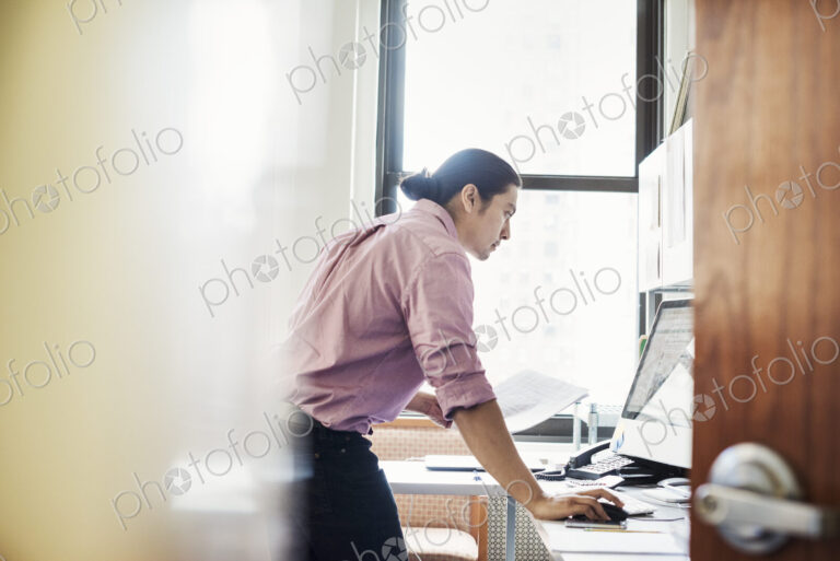 A man standing in his office leaning on the desk, viewed through an open door.