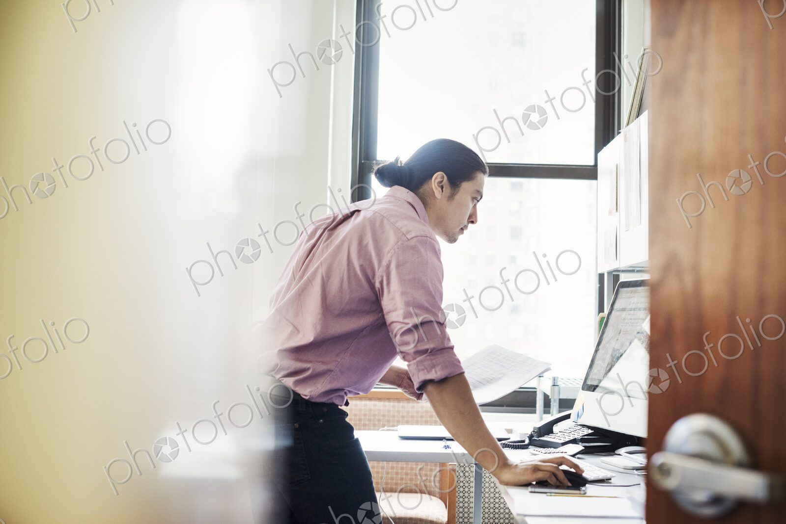 A man standing in his office leaning on the desk, viewed through an open door.