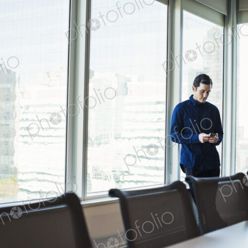 A man standing next to a table in a meeting room looking at a cellphone. A man standing next to a table in a meeting room looking at a cellphone.