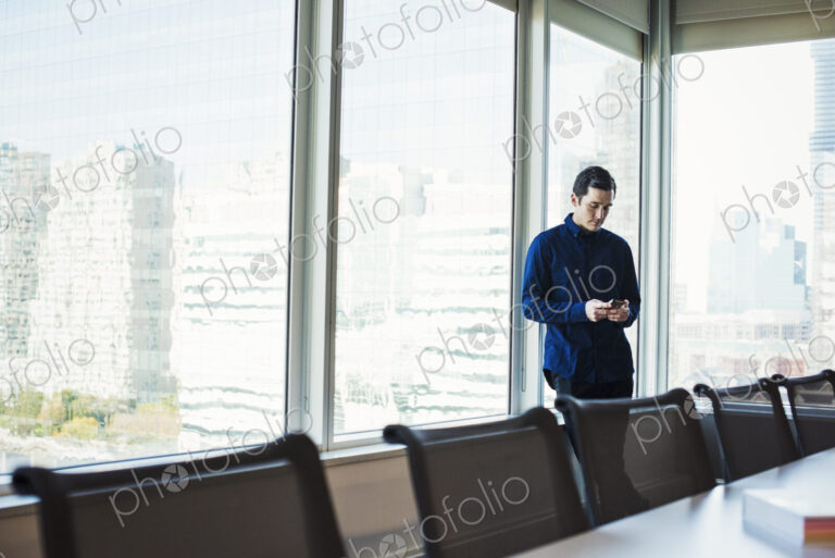 A man standing next to a table in a meeting room looking at a cellphone.