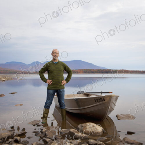 A man standing on a lake shore beside a small rowing boat.