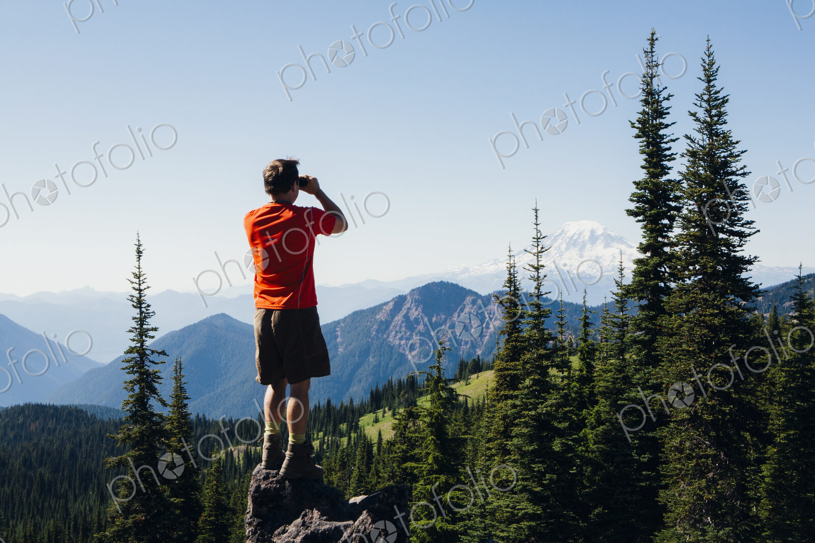 A man standing on a mountain ridge, taking a photograph of the landscape.