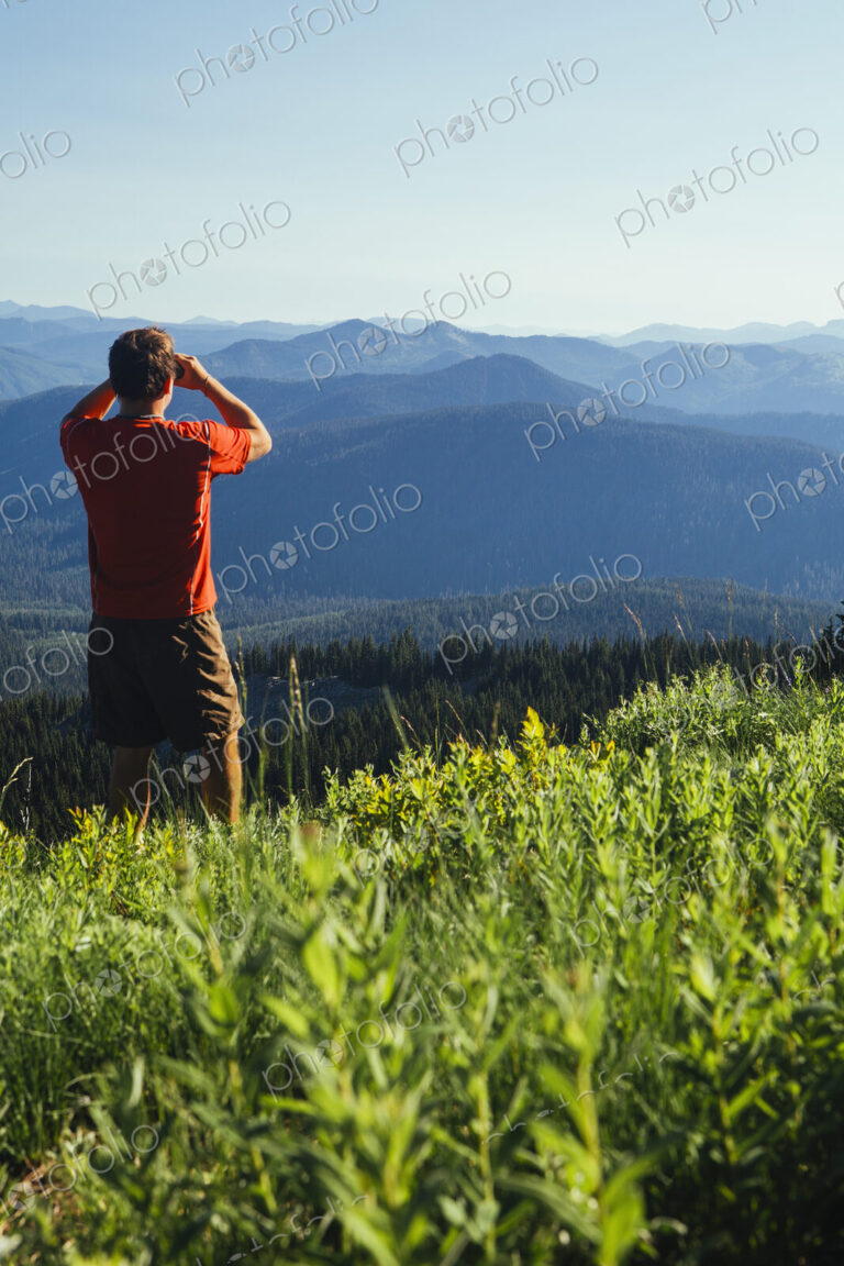 A man standing on a mountain ridge, taking a photograph of the landscape and forested…
