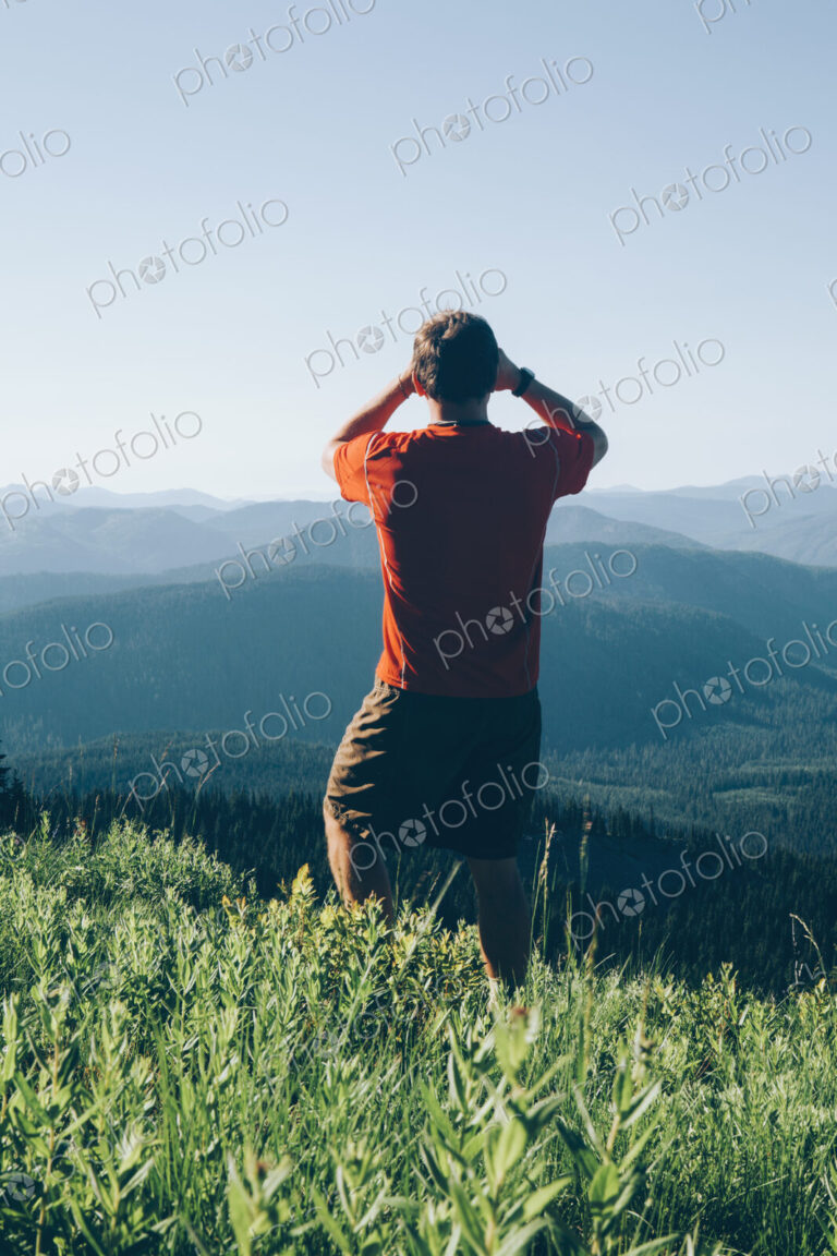 A man standing on a mountain ridge, taking a photograph of the landscape and forested…