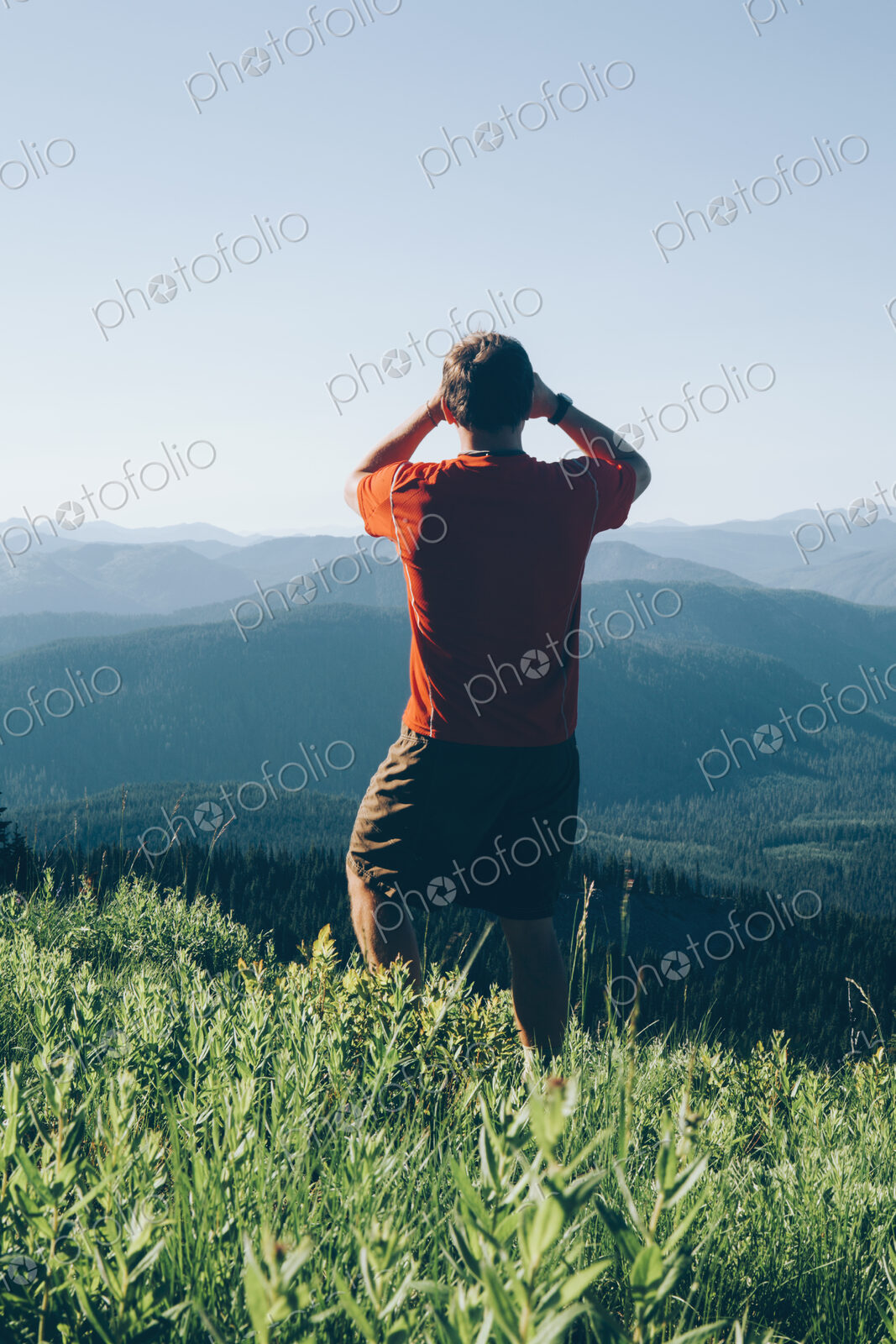 A man standing on a mountain ridge, taking a photograph of the landscape and forested…