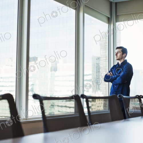 A man standing with arms crossed in a meeting room looking out of a window at an urban… A man standing with arms crossed in a meeting room looking out of a window at an urban…