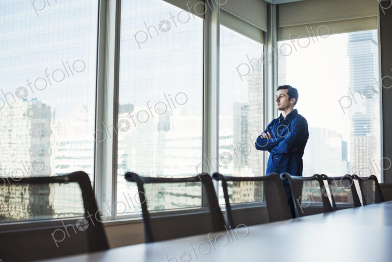 A man standing with arms crossed in a meeting room looking out of a window at an urban…